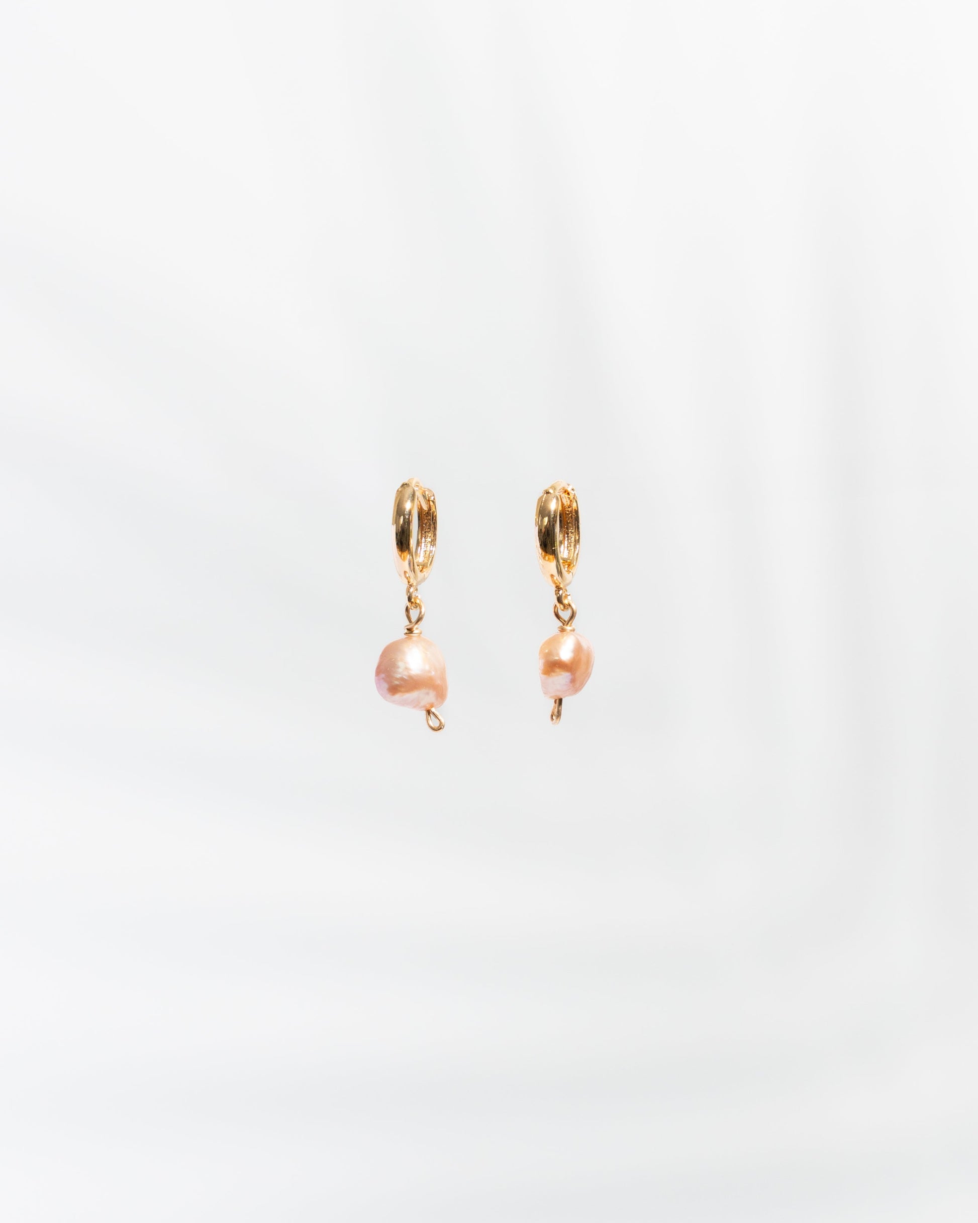 A pair of gold-colored huggie earrings with peach-toned pearls attached, displayed against a white background.