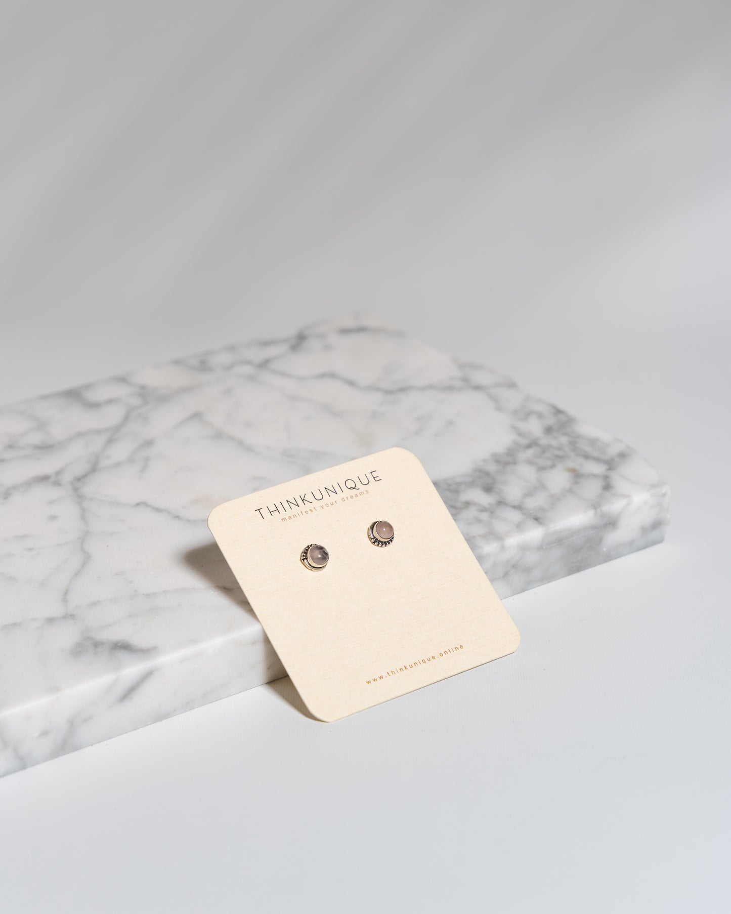 A pair of Mystic Gray Agate stud earrings on a beige card, placed on a marble surface.
